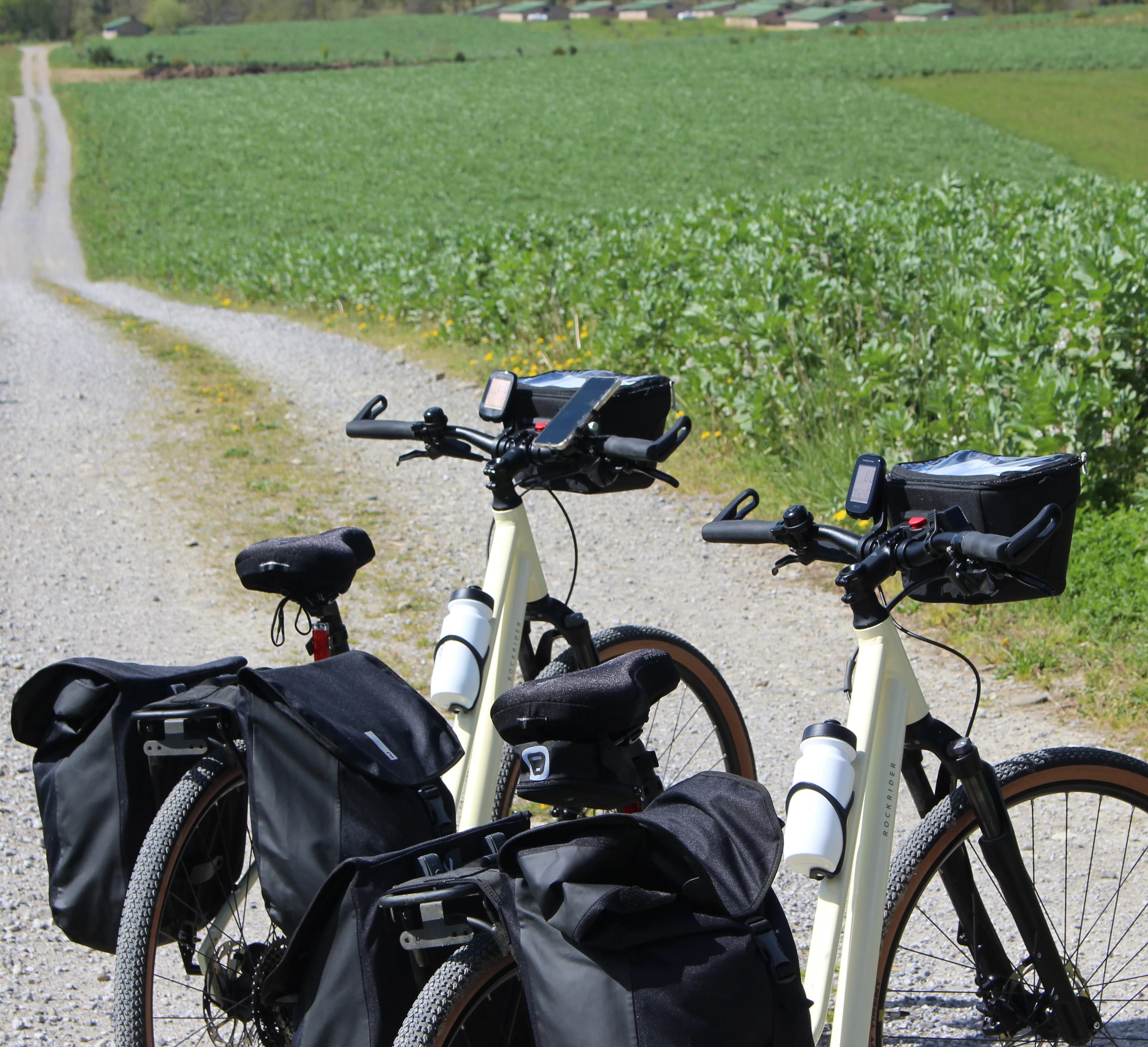 Deux vélos de voyage blancs équipés de sacoches noires, de porte-bouteilles avec gourdes et de supports pour smartphone, stationnés sur un chemin de gravier rural.
Location de vélo de voyage avec l'agence The Family Bike.