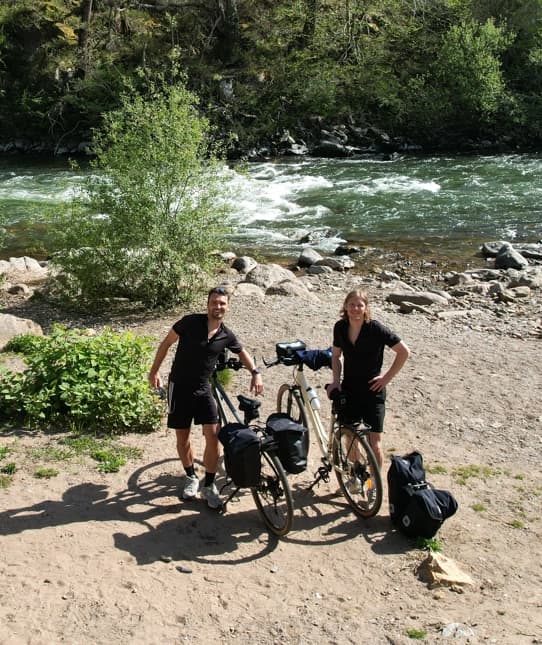 Deux voyageurs à vélo en tenue de sport noire posant avec leurs vélos chargés de sacs près d'une rivière entourée de verdure dans le Pays Basque.
Séjours à vélo clé en main de The Family Bike.